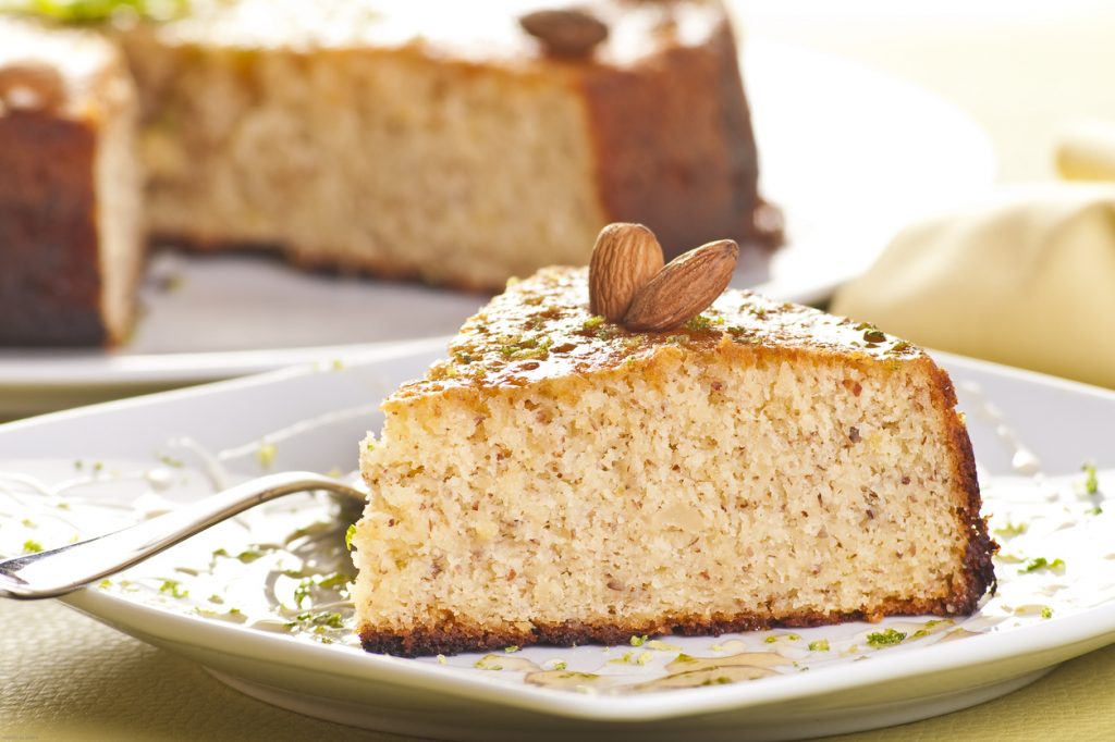 Nahaufnahme eines saftigen Mandelkuchen-Stücks mit goldbrauner Kruste und zwei ganzen Mandeln auf einem Teller.