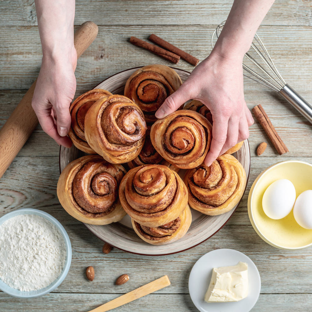 Hände arrangieren goldbraune Zimtschnecken auf einer Servierplatte, umgeben von Mehl, Butter, Eiern, Zimtstangen und Backutensilien auf Holztisch.