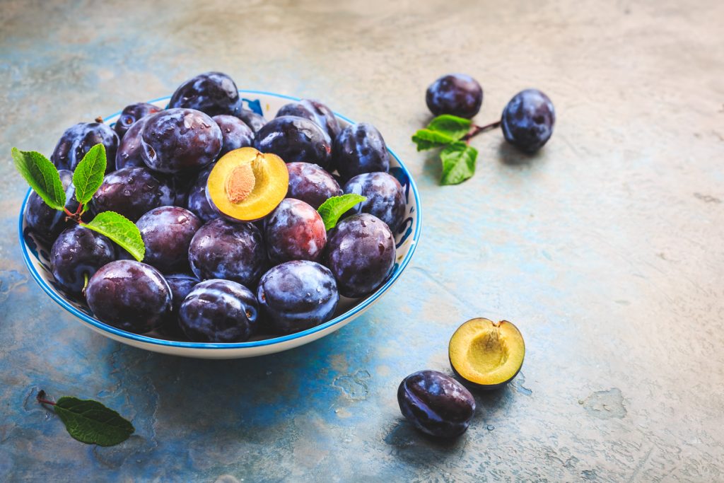 Blauviolette Pflaumen liegen in einer Schale, daneben halbe Pflaumen auf hellem Stein.
