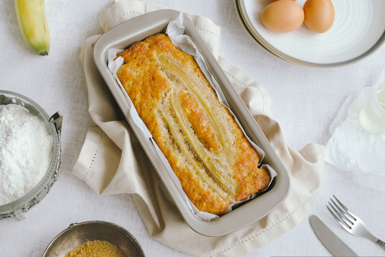 Frisch gebackenes Bananenbrot mit längs aufgelegter Banane in der Kastenform, umgeben von Eiern, Mehl und Zucker.