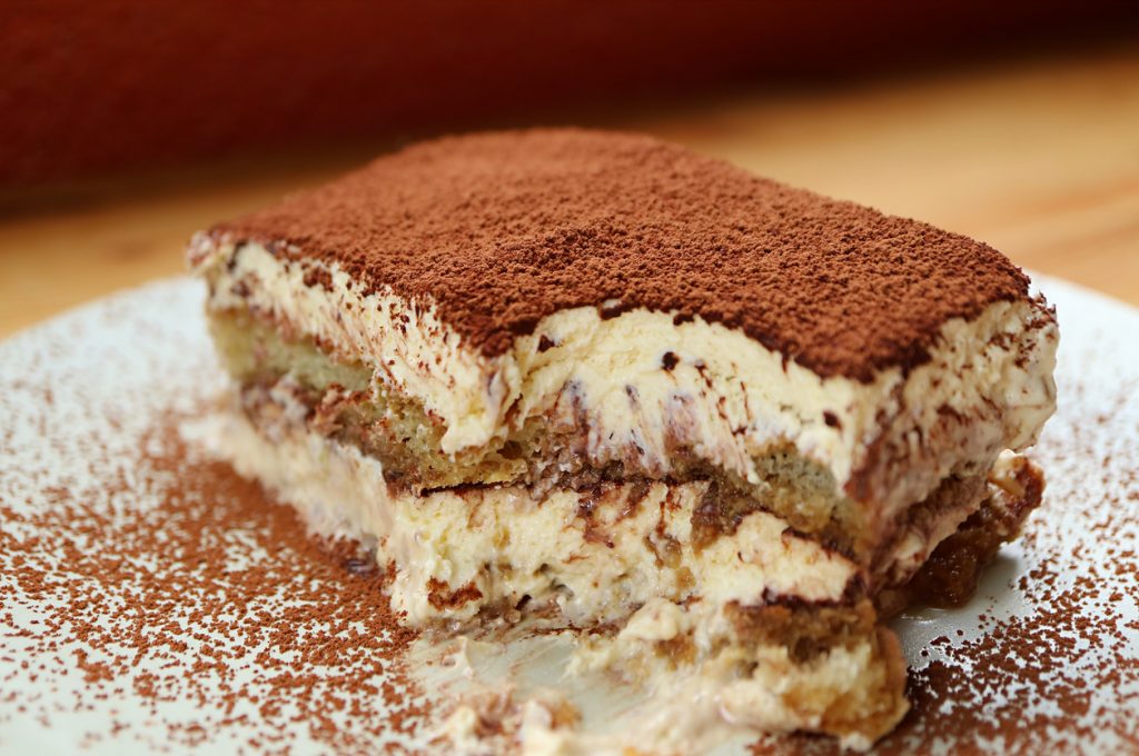 Nahaufnahme eines angeschnittenen Tiramisu-Stücks auf weißem Teller, dick mit Kakaopulver bestäubt; sichtbare Schichten aus Mascarponecreme und espressogetränkten Löffelbiskuits.