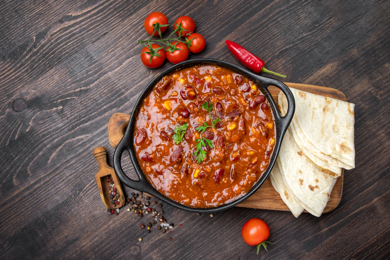Schwarzer Topf mit Chili con Carne, Tortillas und Tomaten auf dunklem Holztisch.