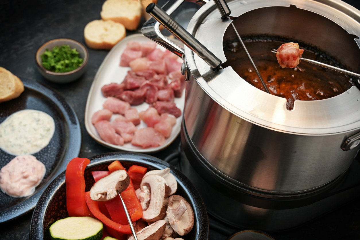 Edelstahl-Fonduetopf mit Brühe und Spießen, daneben Fleischwürfel, Gemüse und Saucen.
