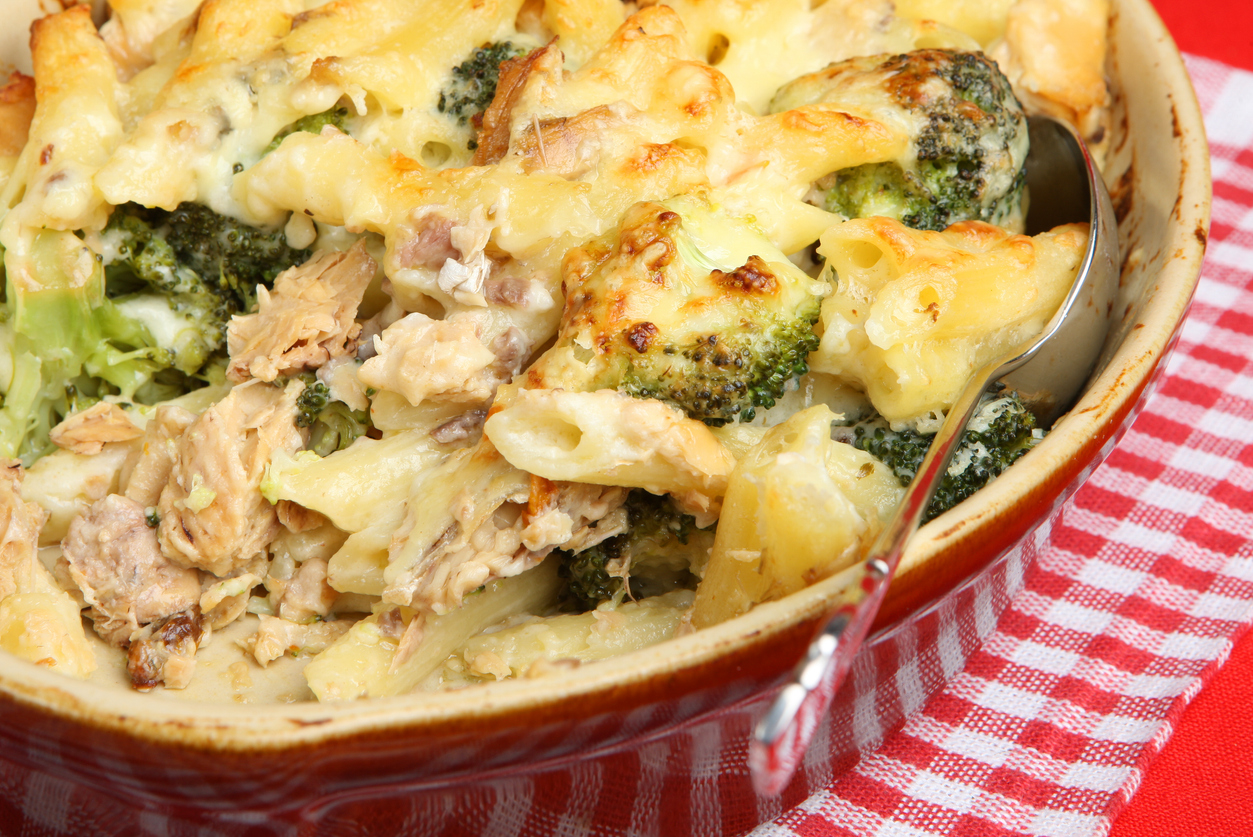 Nahaufnahme eines goldbraun überbackenen Rigatoni-Auflaufs mit Lachs und Brokkoli in cremiger Käsesauce in einer Keramikform mit Löffel auf rotem Karo-Tuch.