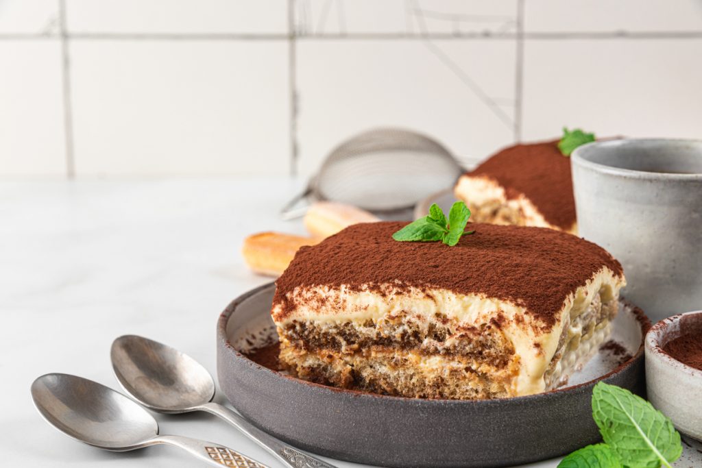 Stück Tiramisu auf grauem Teller, dicht mit Kakaopulver bestäubt und mit Minzblatt garniert; im Hintergrund weiteres Stück, Löffelbiskuits, Sieb und zwei Löffel.