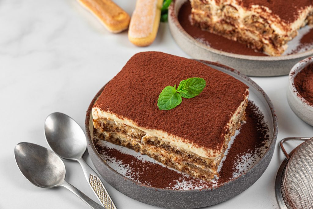 Rechteckiges Stück Tiramisu auf grauem Teller, Oberfläche dicht mit Kakao bestäubt und mit frischem Minzblatt garniert; sichtbare Schichten aus Mascarponecreme und espressogetränkten Löffelbiskuits, daneben zwei Löffel.