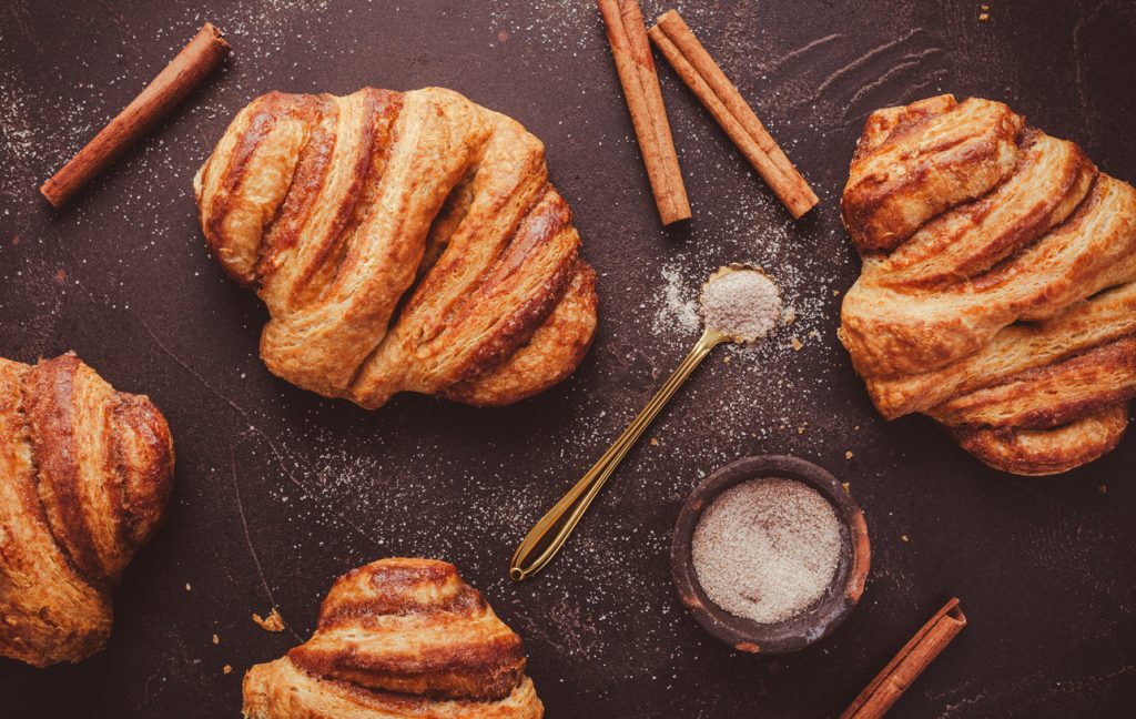 Mehrere goldbraune Franzbrötchen liegen mit Zimtstangen und Zucker auf dunkler Fläche.