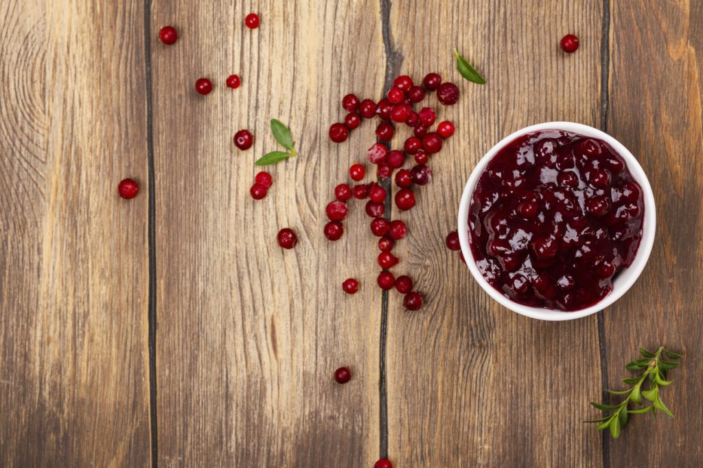 Weiße Schale mit stückiger Cranberrysauce neben verstreuten Cranberries und Grün auf Holztisch.
