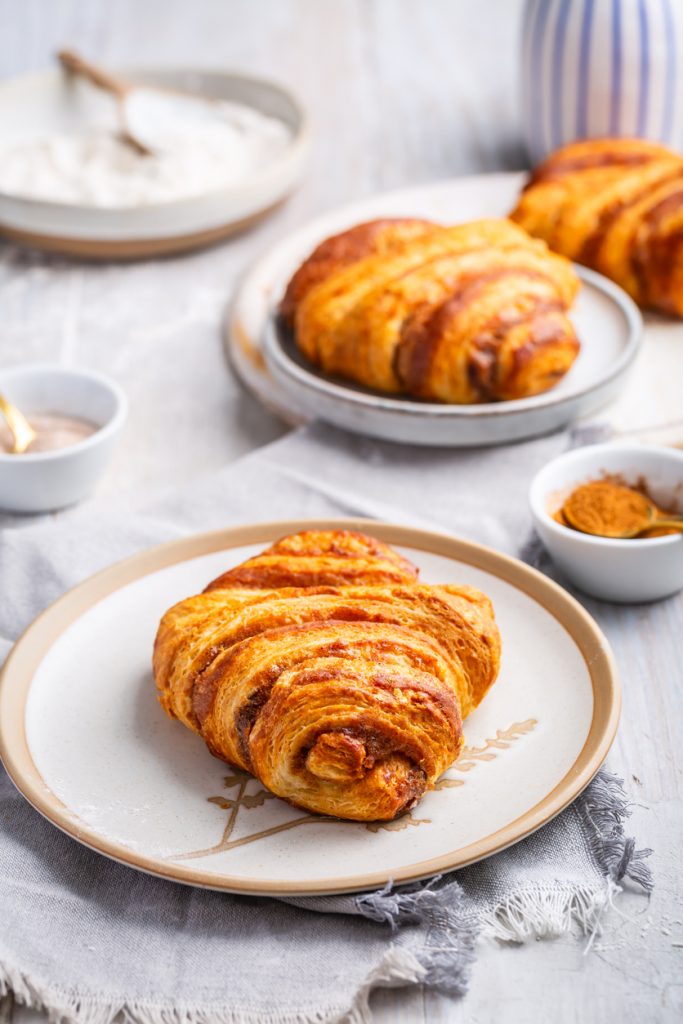 Golden gebackenes Franzbrötchen mit Zimtschichten auf einem Teller, weitere Stücke im Hintergrund.
