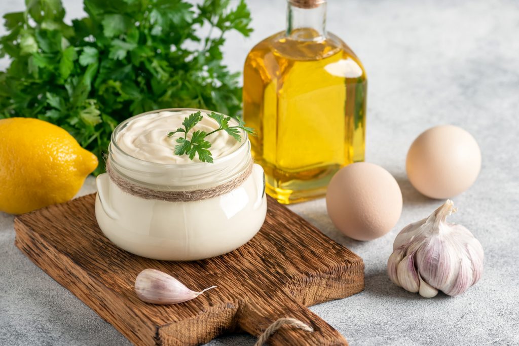 Ein Glas Aioli steht auf einem Holzbrett neben Knoblauch, Eiern, Zitrone und Olivenöl.