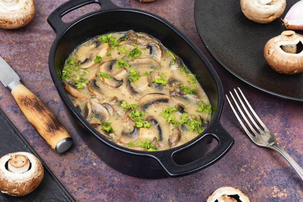 Schwarze Auflaufform mit cremiger Champignonsauce, deutlich sichtbaren Pilzscheiben und Petersilienstreu, daneben Besteck und braune Champignons.