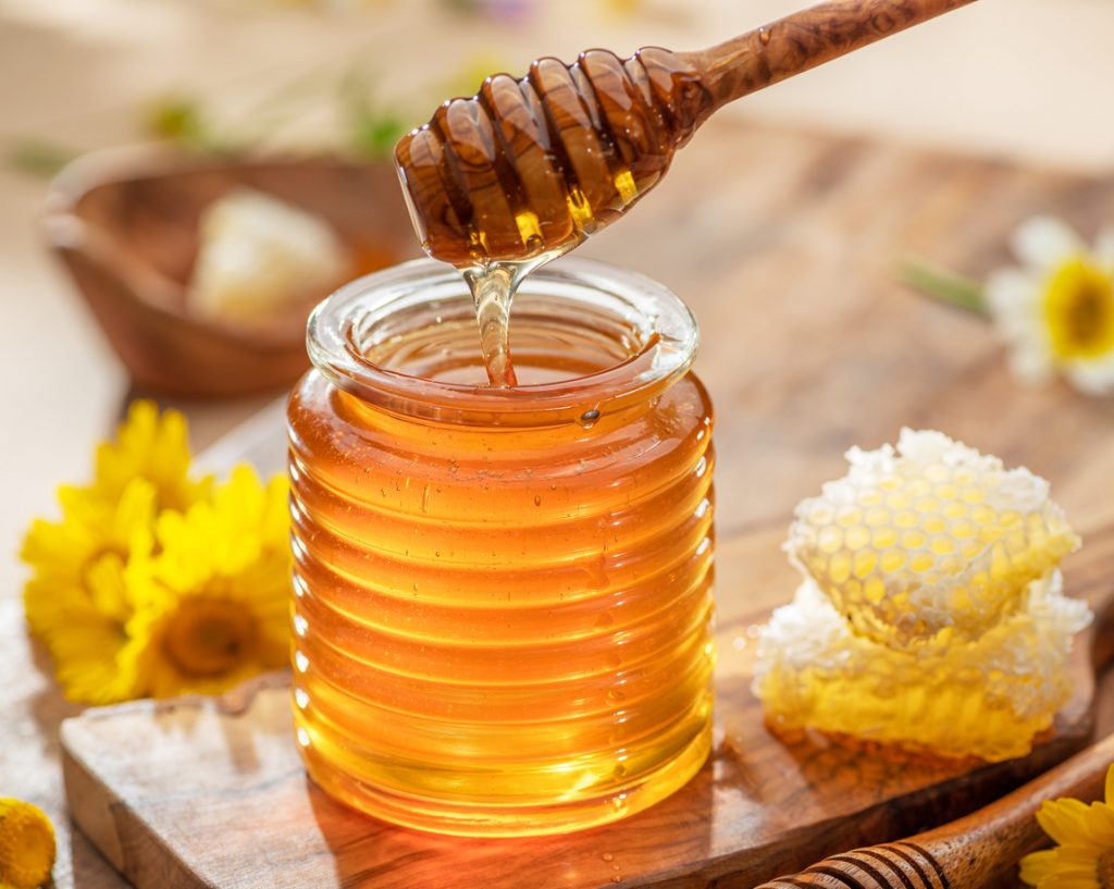 Goldener Honig tropft vom Honiglöffel in ein geriffeltes Glas neben Waben.