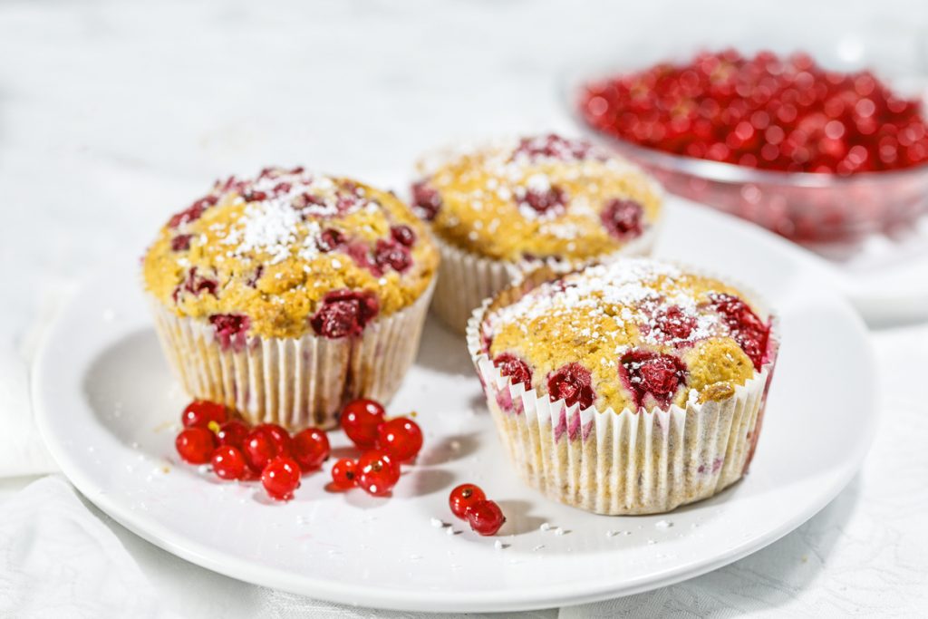 Drei goldbraune Johannisbeer-Muffins auf weißem Teller, mit roten Beeren und Puderzucker bestäubt, daneben lose Johannisbeeren.