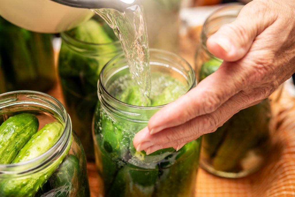 Nahaufnahme: Eine Hand hält ein Glas mit kleinen Gurken, während klare Salzlake aus einem Topf hineingegossen wird.