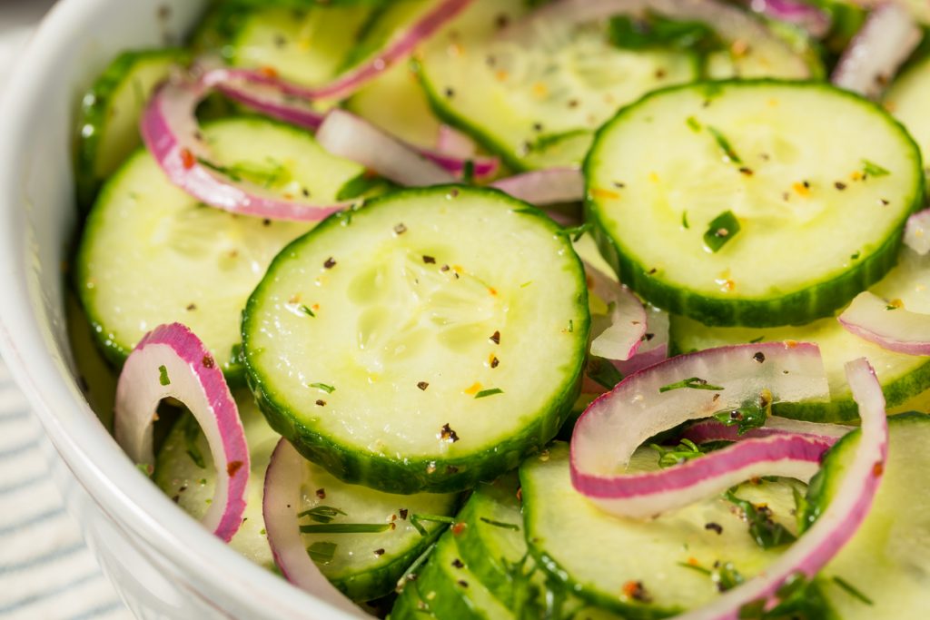 Nahaufnahme von Gurkenscheiben mit roten Zwiebelringen, Dill und Pfeffer in leichter Vinaigrette in einer weißen Schüssel.