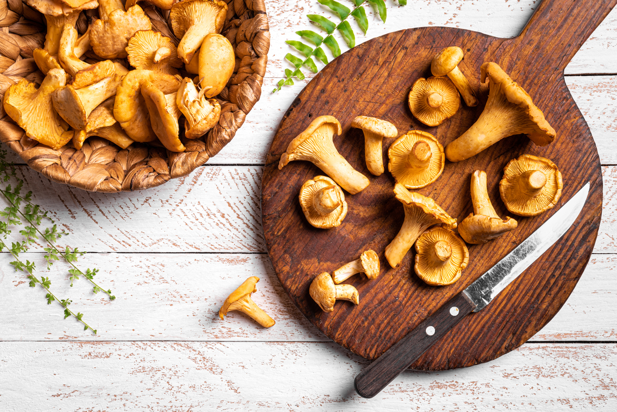 Frische Pfifferlinge auf rundem Holzbrett mit Messer, daneben ein Korb auf weißem Holztisch.
