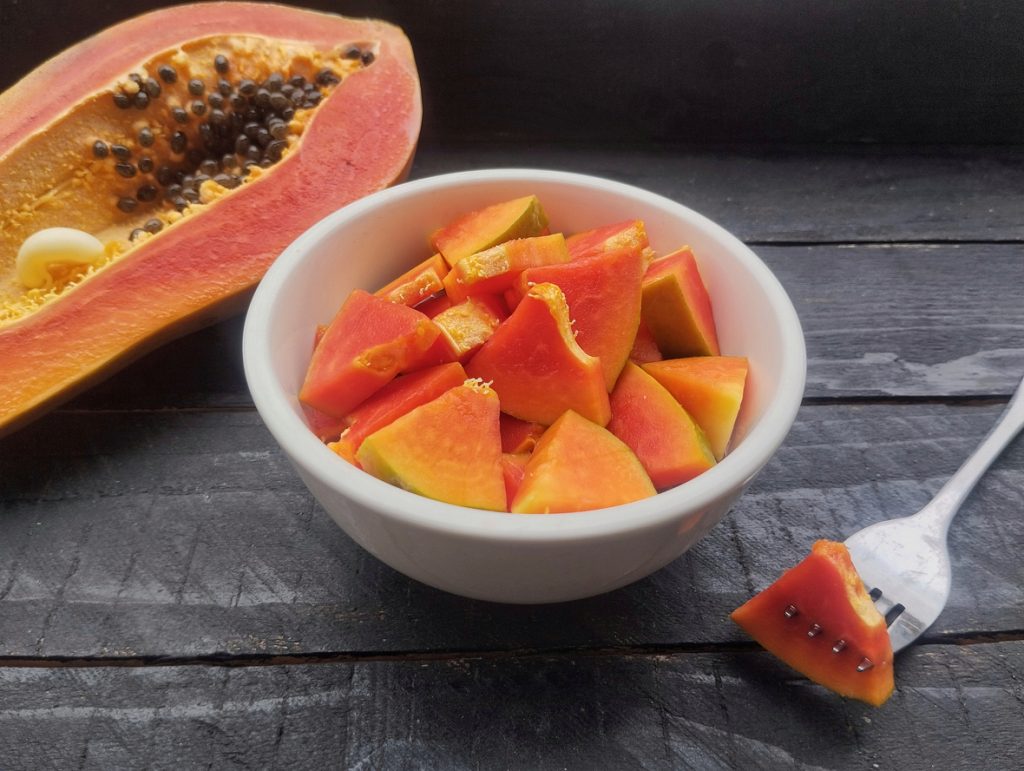 Weiße Schüssel mit gewürfelter reifer Papaya, daneben eine halbierte Papaya mit Kernen und eine Gabel mit einem Stück