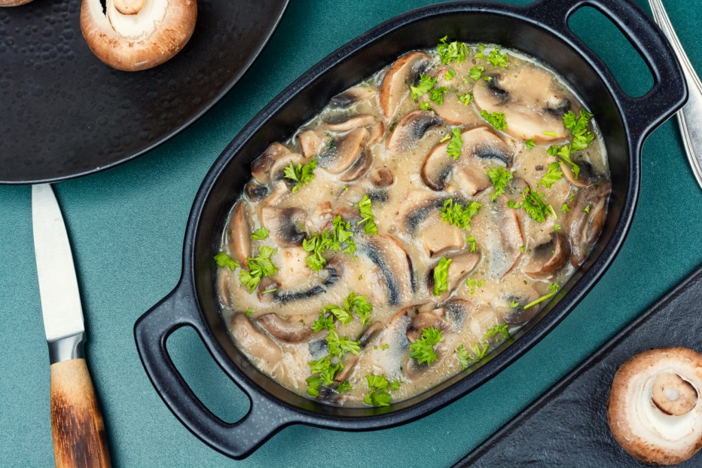 Ovale schwarze Form mit sahniger Champignonsauce, sichtbaren Pilzscheiben und Petersilienstreu auf grünem Untergrund, daneben Messer und Champignons.