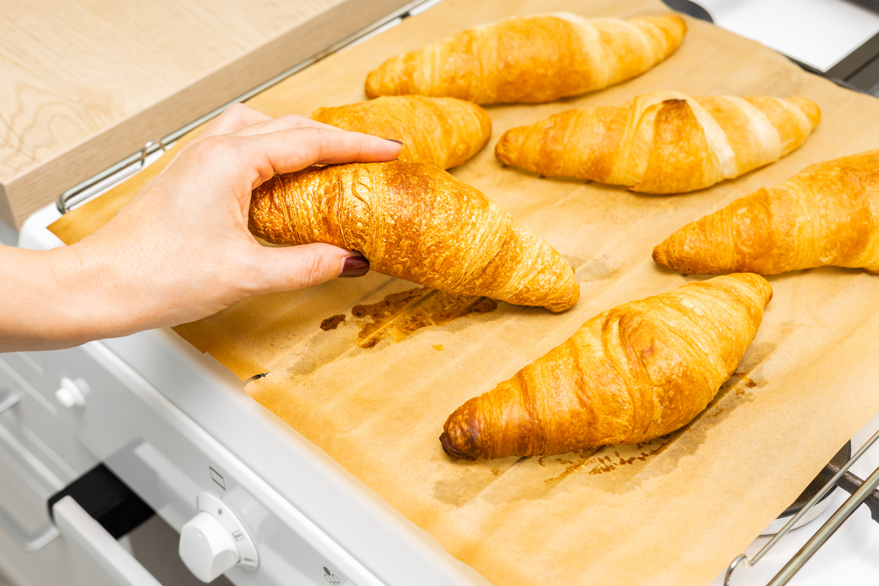 Hand nimmt ein goldbraunes Buttercroissant von einem mit Backpapier belegten Blech aus dem Ofen.