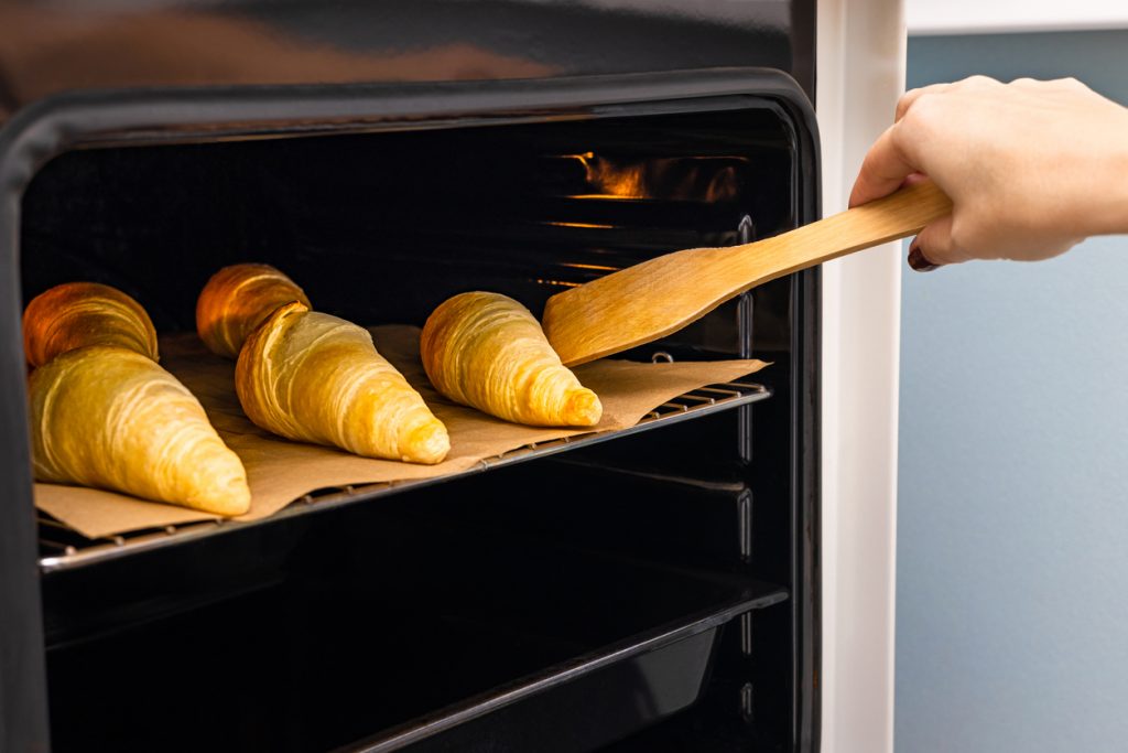 Hand richtet mit einem Holzspatel goldene Croissants auf Backpapier im heißen Ofen aus.