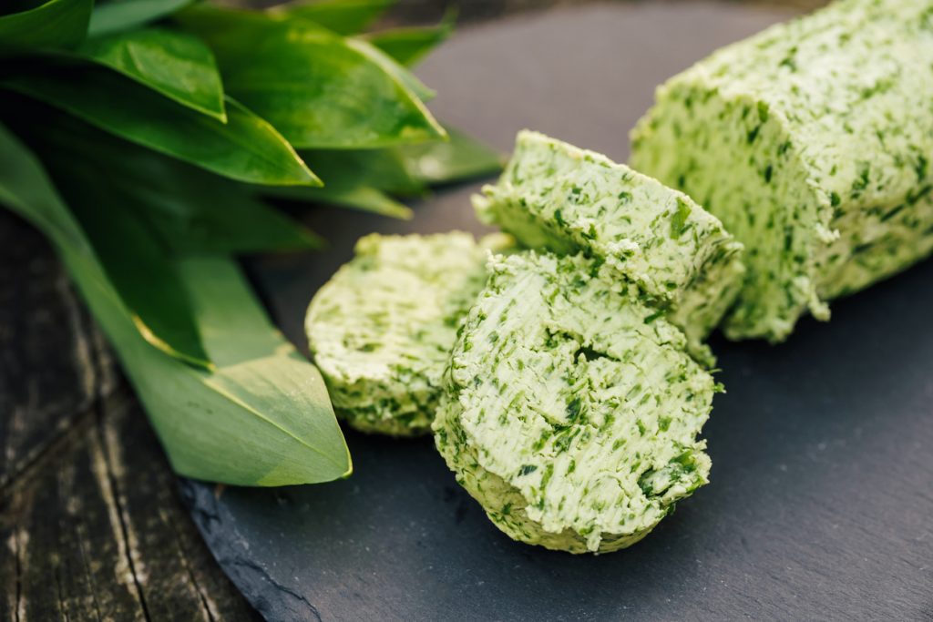 Nahaufnahme von in Scheiben geschnittener Bärlauchbutter auf dunkler Schieferplatte mit frischen Bärlauchblättern.