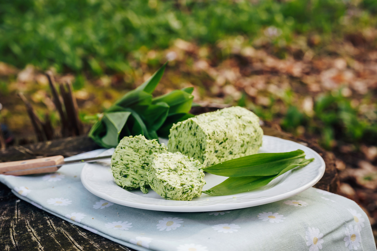 Aufgeschnittene Rolle Bärlauchbutter mit frischen Bärlauchblättern auf weißem Teller im Freien.