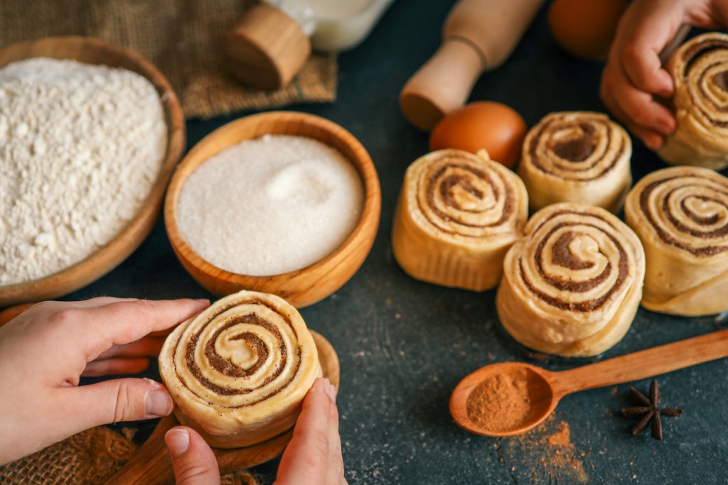 Hände formen ungebackene Zimtschnecken, umgeben von Mehl- und Zuckerschalen, Eiern, Milch und einem Löffel Zimt.