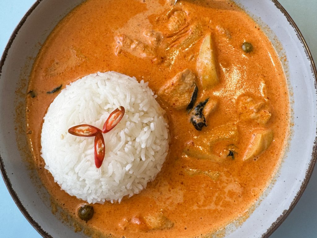 Nahaufnahme eines tiefen Tellers mit cremigem rotem Kokosnuss-Thai-Curry, zartem Hühnchen und geformtem Jasminreis, garniert mit Chilischeiben.