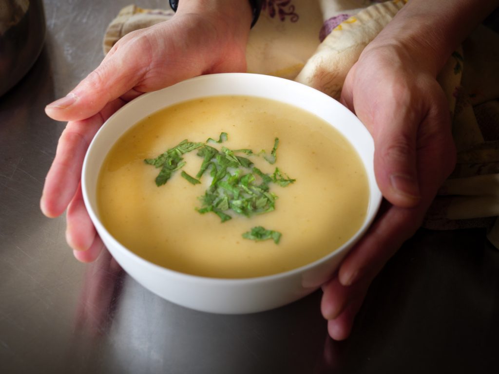 Weiße Schale mit cremiger Pastinakensuppe auf Kokosmilchbasis, in Händen gehalten und mit frischen Kräutern bestreut.