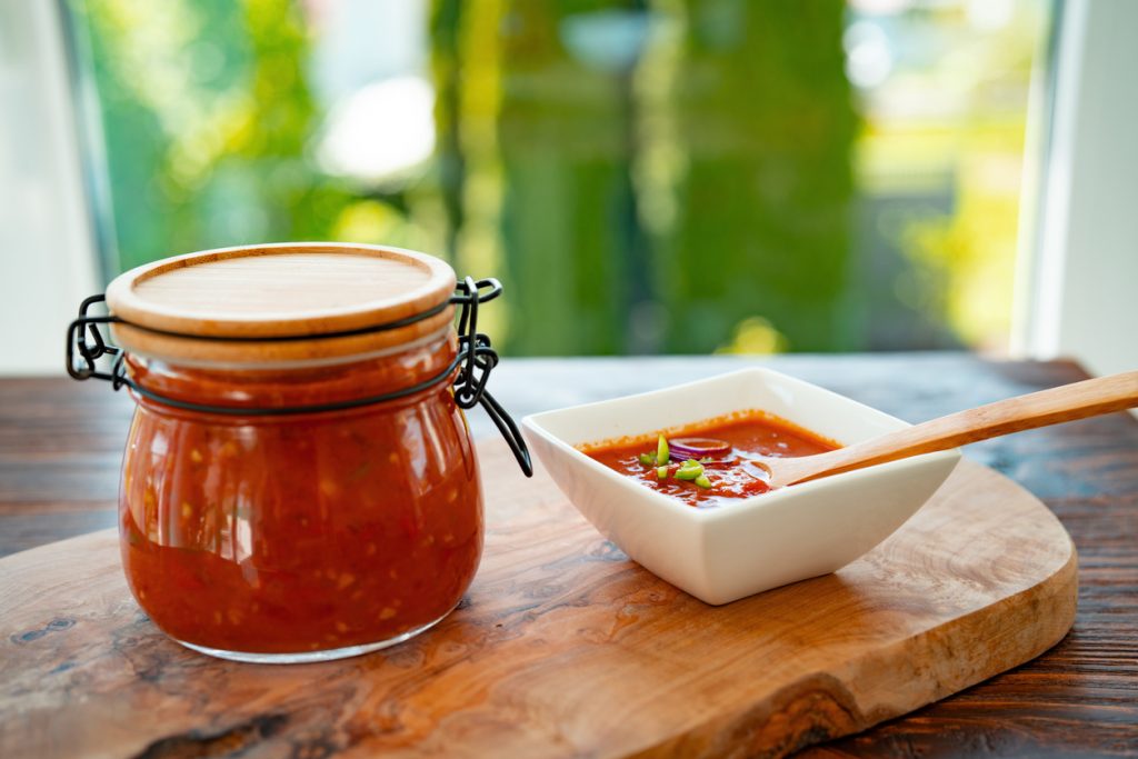 Ein Einmachglas mit roter Salsa steht neben einer weißen Schale mit Salsa, Chilischeiben und Holzlöffel auf einem Holzbrett.