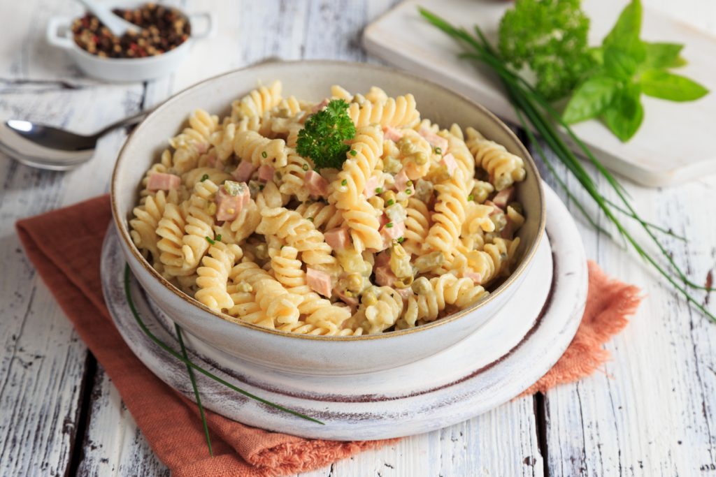 Schüssel mit Fusilli-Nudelsalat in cremigem Dressing, gemischt mit Schinkenwürfeln, Erbsen und Schnittlauch, garniert mit Petersilie