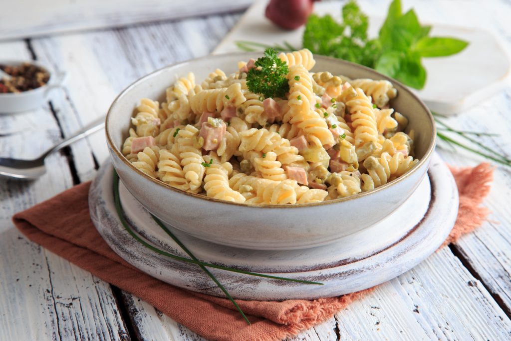 Graue Schüssel mit cremigem Fusilli-Nudelsalat, gemischt mit Schinkenwürfeln, Erbsen und Schnittlauch, garniert mit Petersilie auf rustikalem Tisch