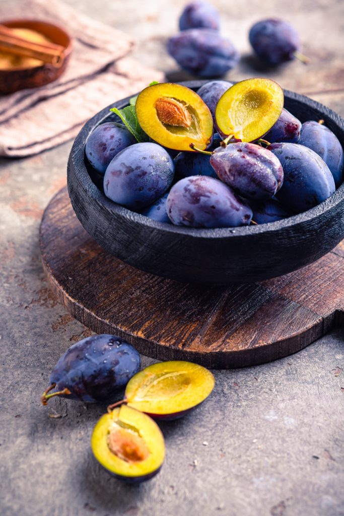 Schwarze Schale mit reifen Pflaumen auf Holzbrett, vorn halbierte Frucht.