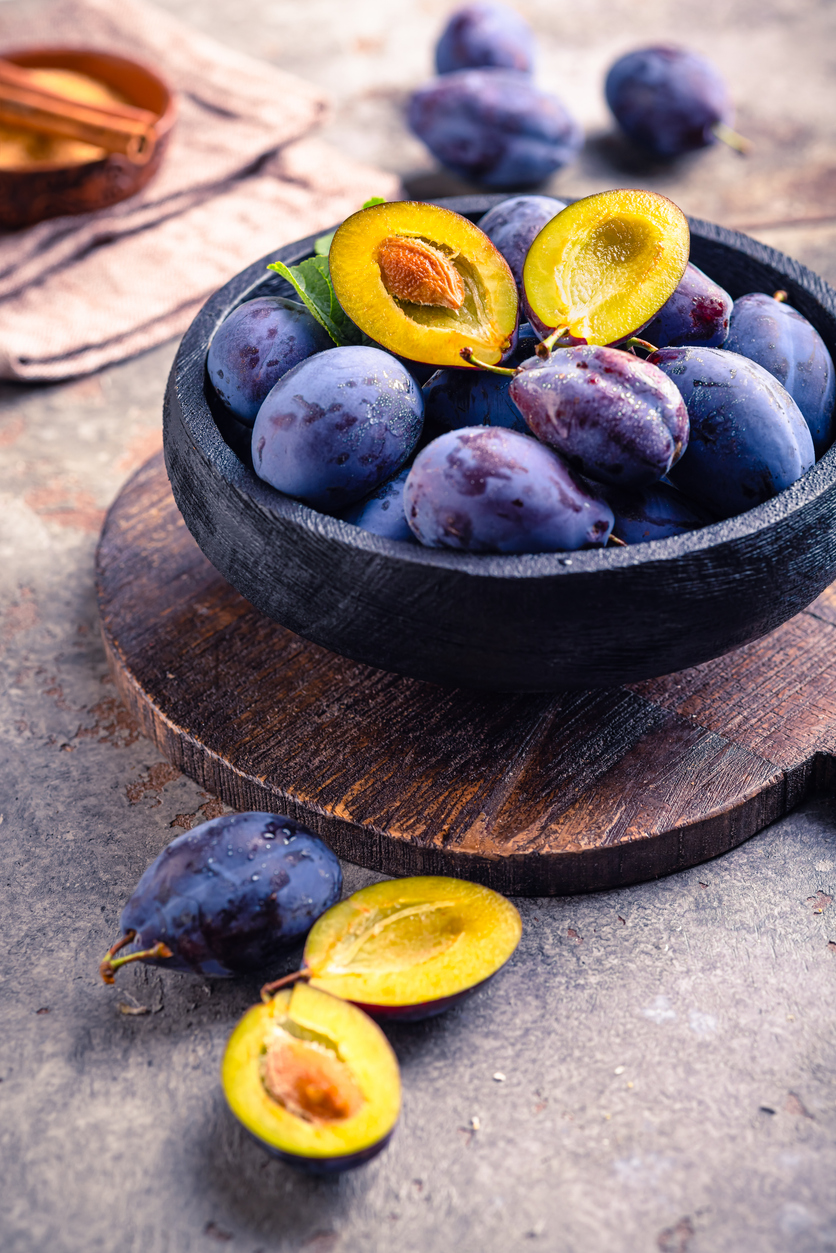 Schwarze Schale mit reifen Pflaumen auf Holzbrett, vorn halbierte Frucht.