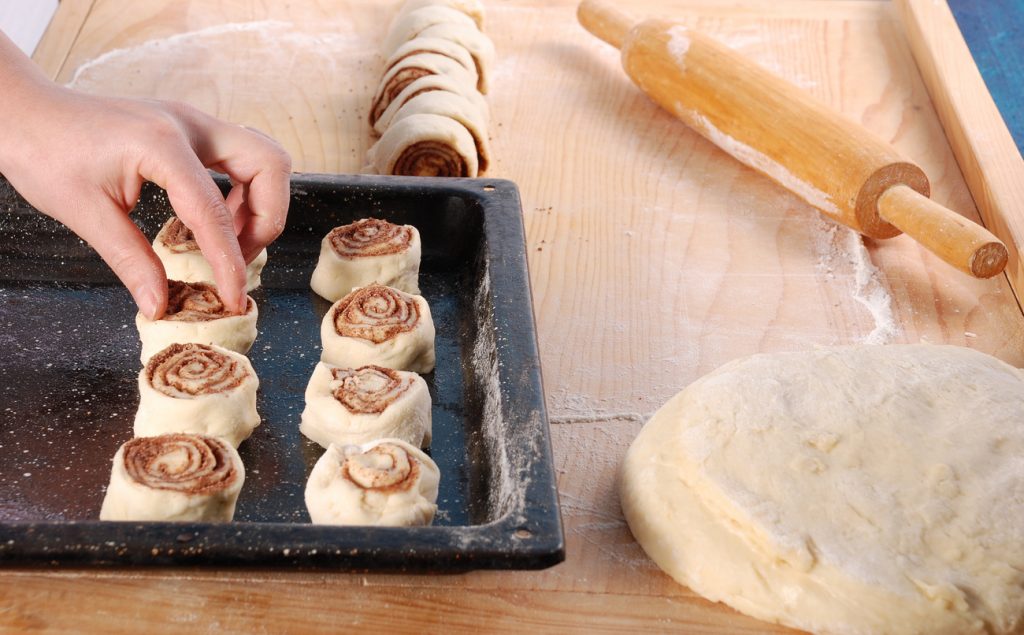 Hand setzt ungebackene Zimtschnecken in ein Backblech, daneben liegen Teigrolle, Nudelholz und bemehlte Arbeitsfläche.