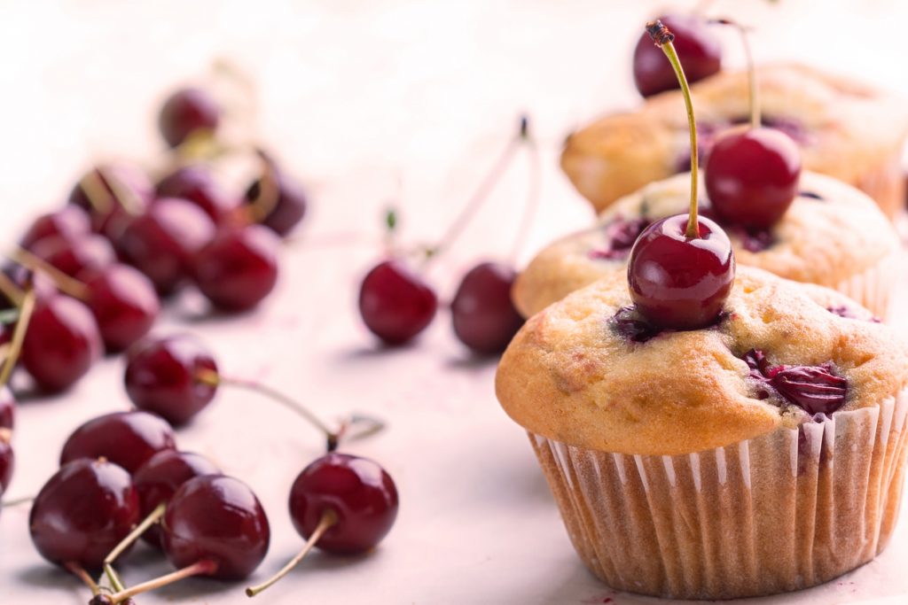 Goldbraune Kirsch-Muffins mit ganzen Kirschen obenauf, umgeben von verstreuten roten Kirschen auf hellem Untergrund.