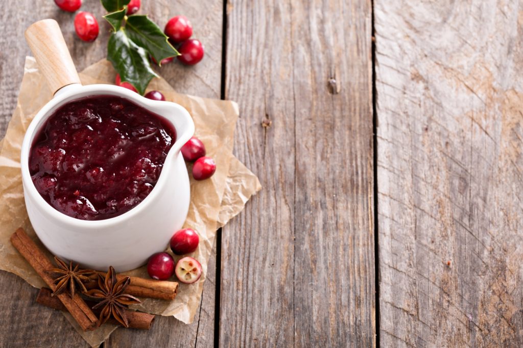 Weiße Sauciere mit dicker Cranberrysauce, umgeben von Cranberries, Zimtstangen und Sternanis auf rustikalem Holztisch.