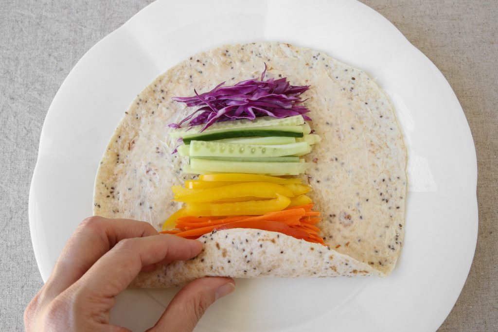 Offene Tortilla mit Frischkäse, belegt in Streifen mit Rotkohl, Gurke, gelber Paprika und Karotte; eine Hand rollt sie an.