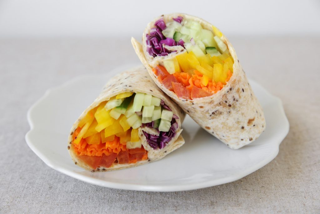 Zwei halbierte Tortilla-Wraps auf weißem Teller, gefüllt mit Rotkohl, Gurke, gelber Paprika, Karotte und Tomate.