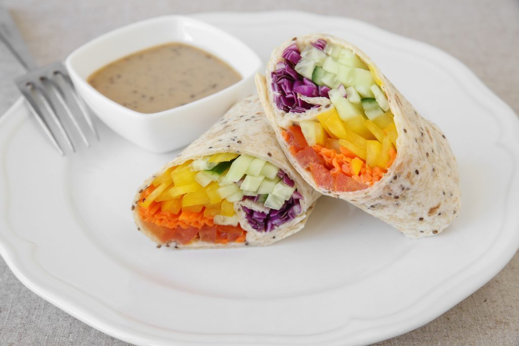 Zwei halbierte Vollkorn-Wraps mit Rotkohl, Gurke, gelber Paprika, Karotte und Tomate auf weißem Teller, dazu eine kleine Dipschale und eine Gabel.
