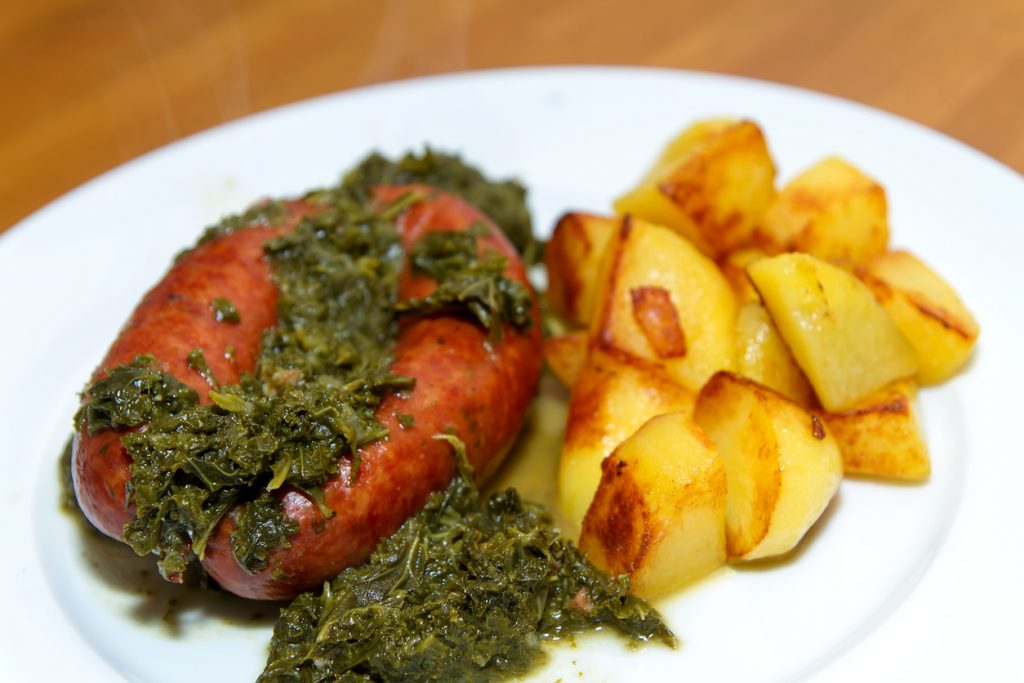 Teller mit gebratener Rauchwurst, bedeckt mit Grünkohl, daneben goldbraune Bratkartoffelwürfel.