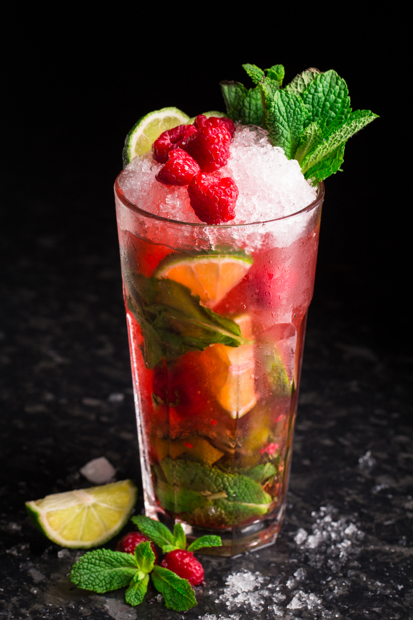 Hoher Cocktail mit Crushed Ice, Limettenstücken, Minze und Himbeeren in einem Glas auf dunkler Fläche.