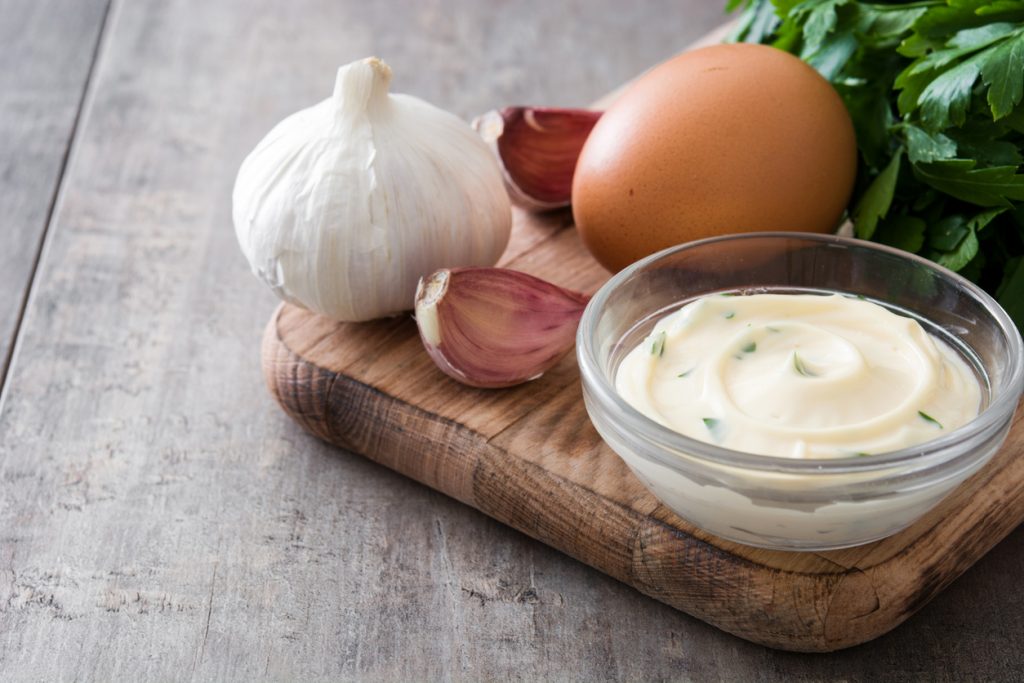 Glasschale mit cremiger Aioli neben Knoblauch, Ei und Petersilie