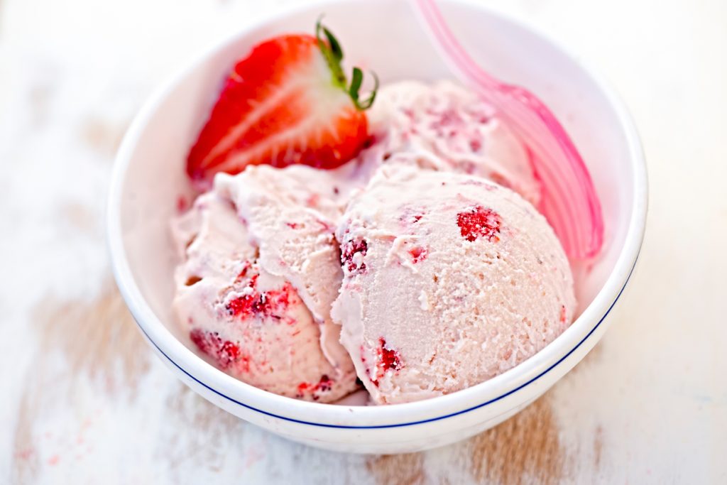 Zwei Kugeln Erdbeereis mit halber Erdbeere und rosa Löffel.