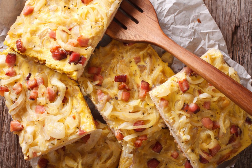 Mehrere Stücke Zwiebelkuchen mit Zwiebelringen und Speck liegen auf Backpapier.