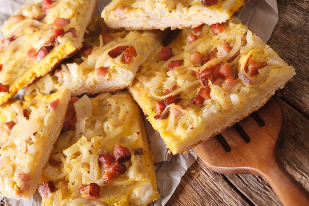Stücke Zwiebelkuchen mit goldbraunen Zwiebeln und Speck auf Holzbrett.