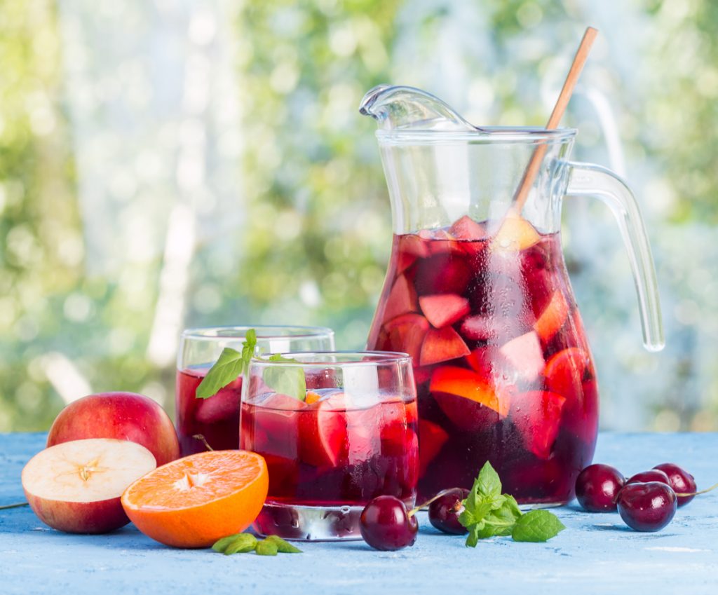 Karaffe und zwei Gläser rote Sangria mit Eiswürfeln und Obststücken, daneben halbierte Orange, Apfel und Kirschen.
