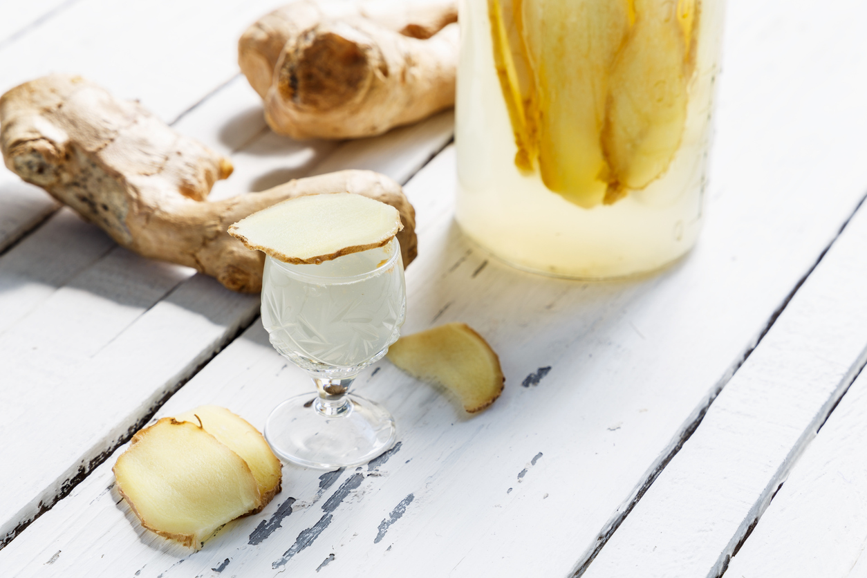 Kleines Likörglas mit hellem Ingwer-Likör und Ingwerscheibe am Rand, daneben Ingwerwurzeln und ein Gefäß mit eingelegten Scheiben auf weißem Holztisch.