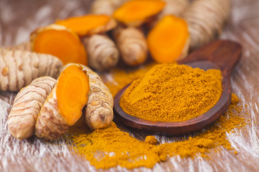 Holzlöffel mit leuchtend orangem Kurkumapulver, umgeben von ganzen und aufgeschnittenen Kurkumawurzeln.