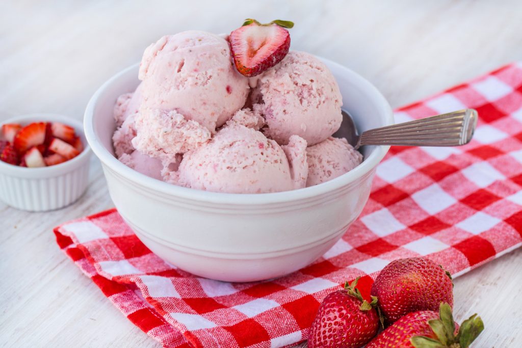 Weiße Schale mit rosa Erdbeereis auf kariertem Tuch neben Erdbeeren.